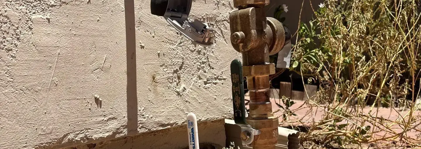 Outdoor irrigation valve and piping with rusted fittings, set against a stucco wall; a black plastic device hangs from the pipe.