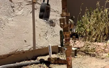 Outdoor irrigation valve and piping with rusted fittings, set against a stucco wall; a black plastic device hangs from the pipe.